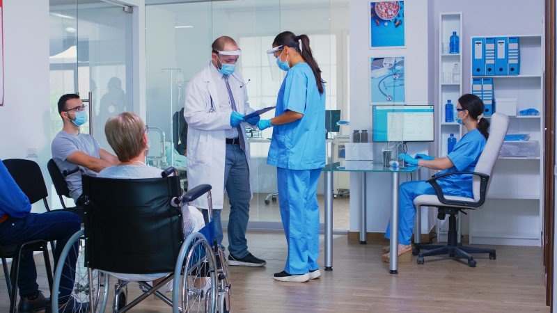 Doctor in a face mask discussing medical information with a nurse while patients, including a woman in a wheelchair, wait in a hospital.