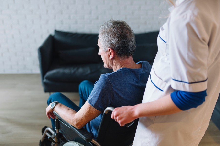 Older man sitting in a wheelchair while a caregiver assists him indoors.