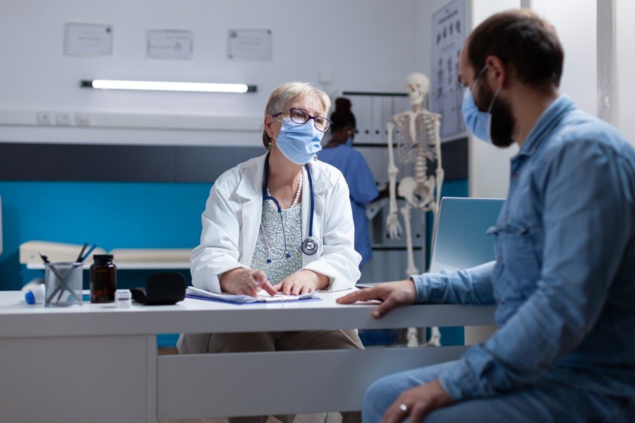 Doctor wearing a face mask consulting with a masked male patient in a medical office.