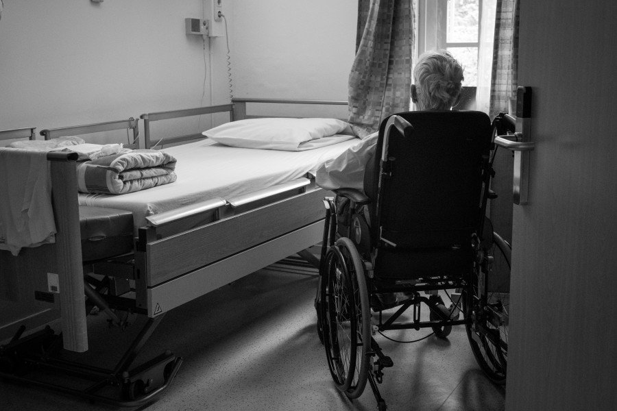 Elderly resident in a wheelchair beside a hospital bed in a Nursing home room.