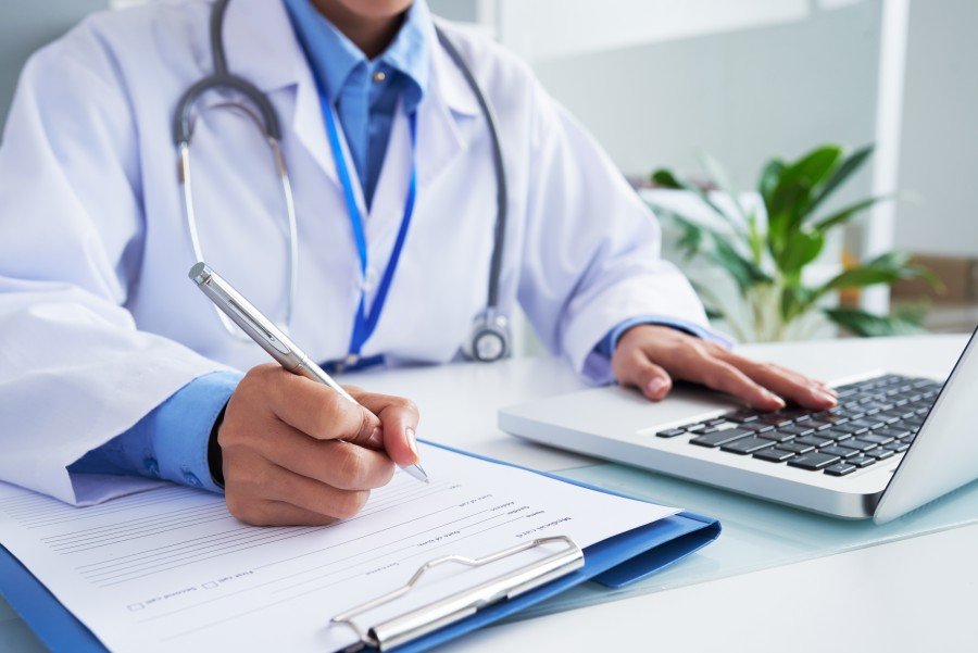 General Physician in a white coat writing on a medical form beside a laptop.