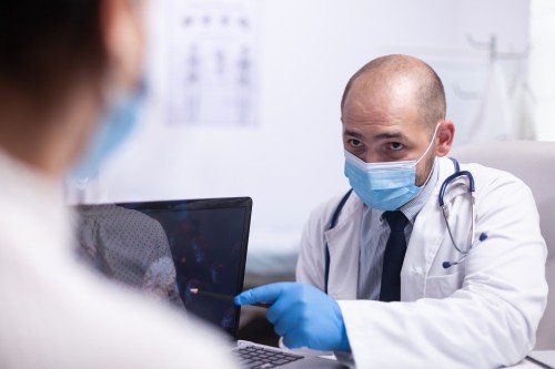 Urologist wearing a surgical mask and blue gloves, discussing medical information with a patient while pointing at a laptop in a clinic